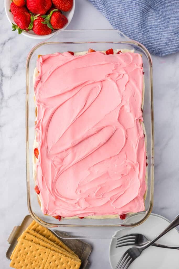 A rectangular glass dish filled with a finished no bake strawberry chocolate eclair cake topped with pink strawberry frosting.
