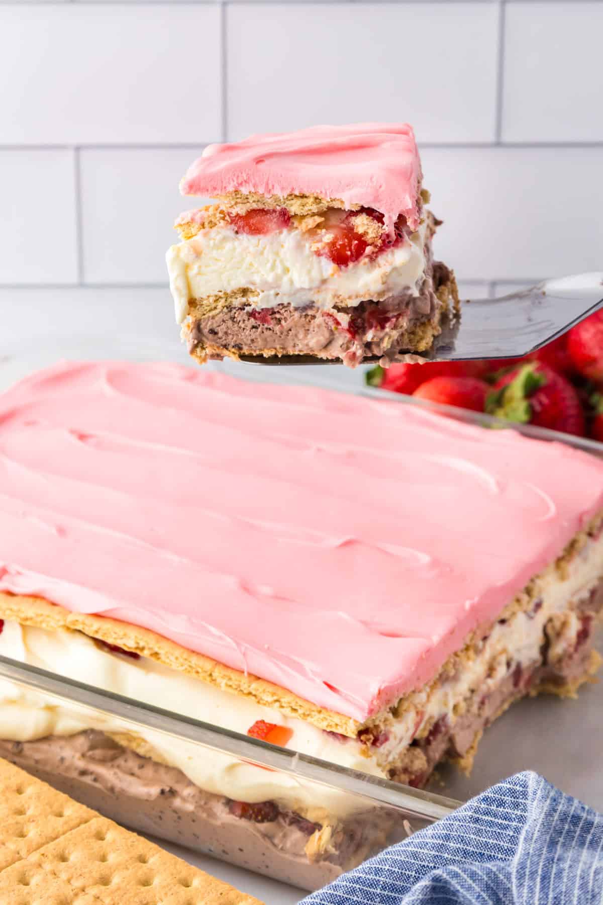 A slice of layered strawberry chocolate eclair cake with pink frosting is lifted from a baking dish full of more of the dessert.