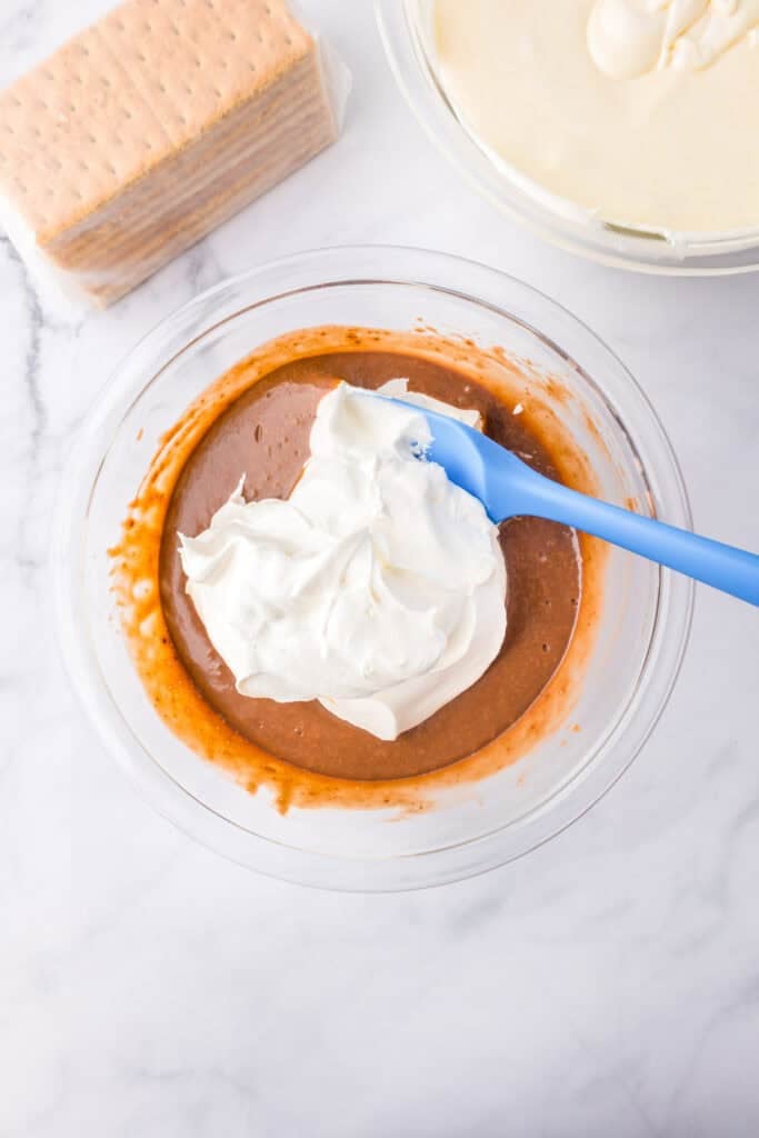 A bowl with Cool Whip being folded into chocolate instant pudding for strawberry chocolate eclair cake with graham crackers and cheesecake pudding nearby.