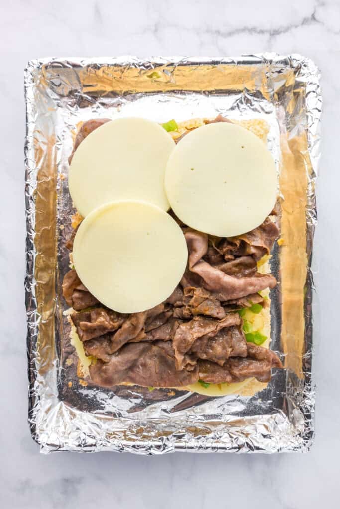 A foil lined pan with the bottom half of Hawaiaan rolls topped with a few slices of provelone cheese, sliced roast beef, peppers and onions for philly cheesesteak sliders.