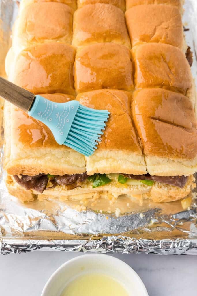 A blue silicone brush spreads melted butter on top of philly cheesesteak slider sandwiches arranged on foil lined pan.