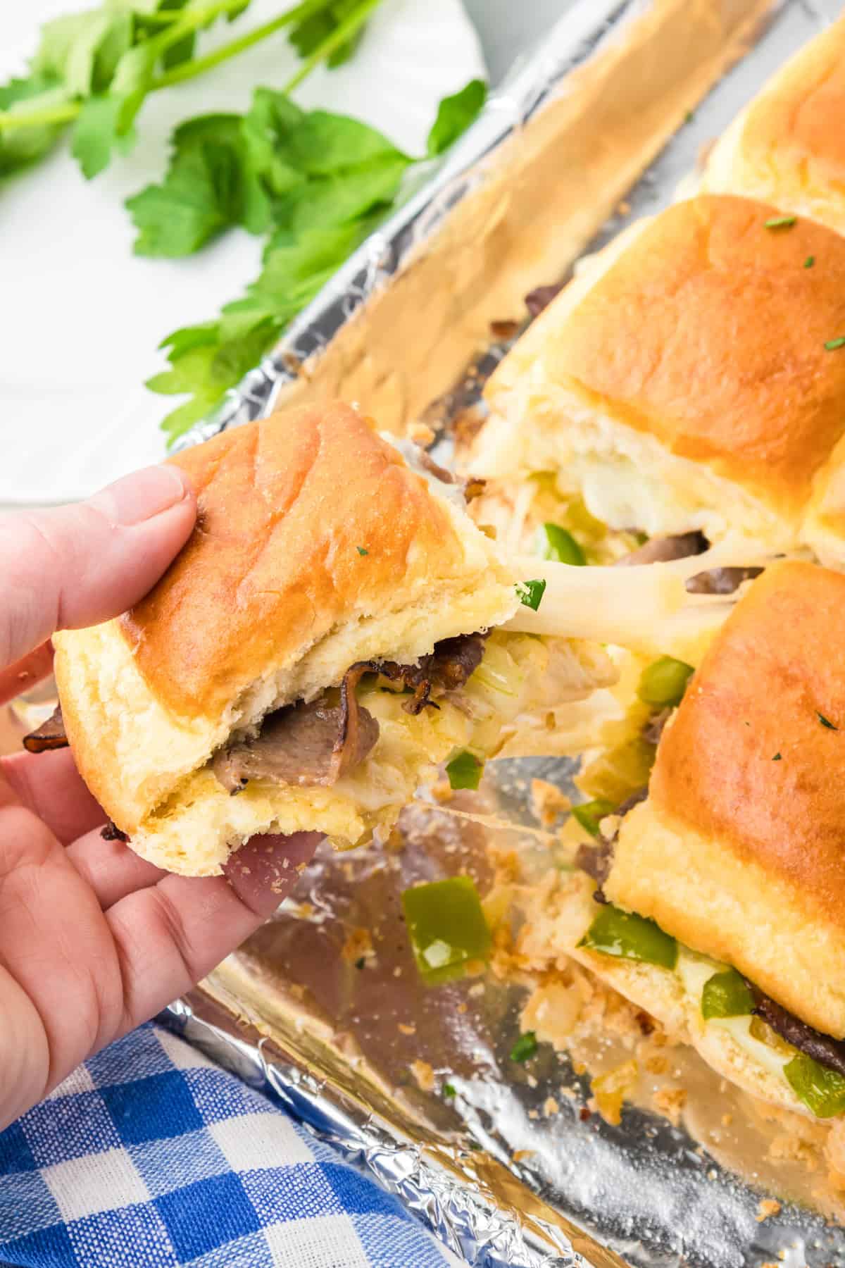 A hand holding a cheesy philly cheesesteak slider sandwich being pulled from a pan full of sliders.