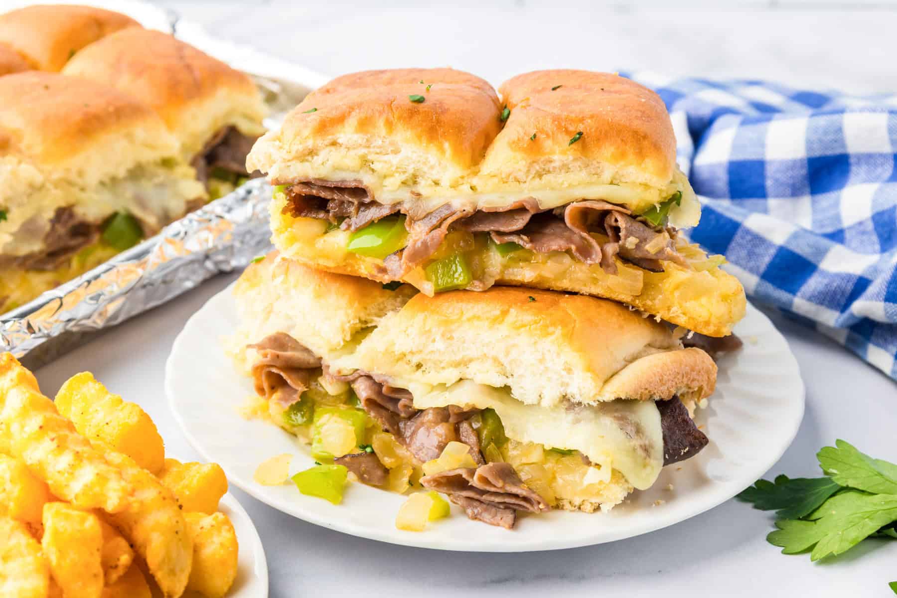 Four philly cheesesteak sliders with beef, cheese, green peppers, and onions are stacked on a white plate.