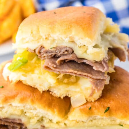 A close-up of three philly cheesesteak sliders with melted cheese, chopped green onions, and onions on Hawaiian rolls.