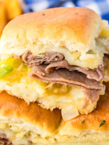 A close-up of three philly cheesesteak sliders with melted cheese, chopped green onions, and onions on Hawaiian rolls.