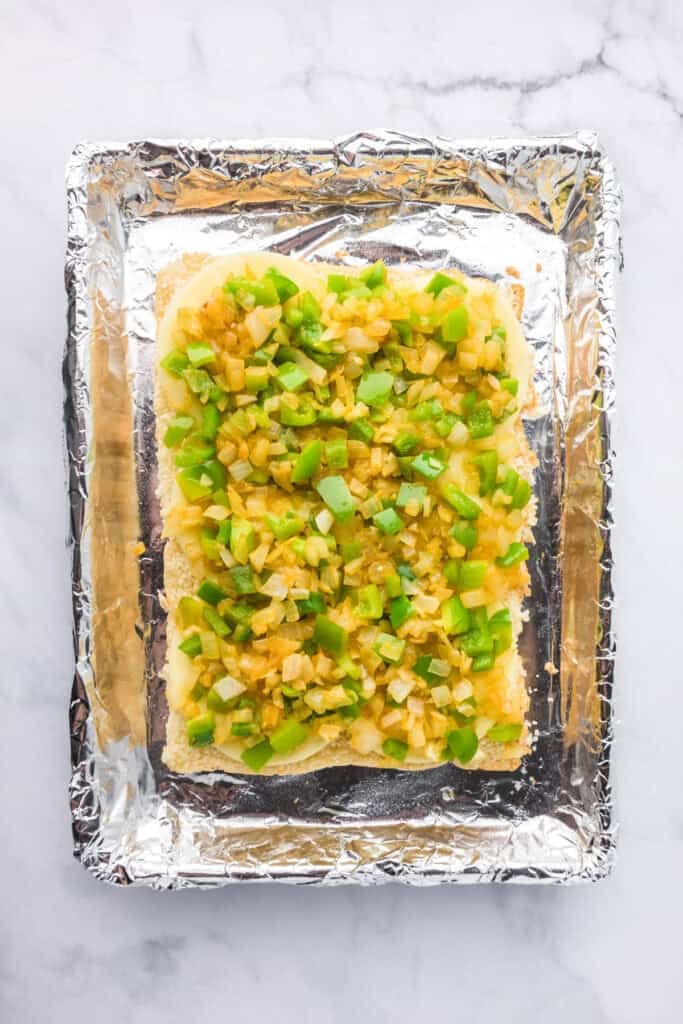 A foil lined pan with the bottom half of Hawaiaan rolls topped with cheese, peppers and onions for philly cheesesteak sliders.