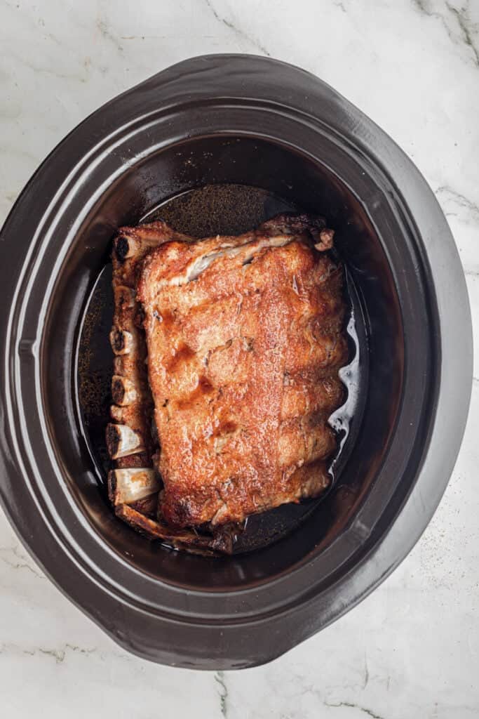 A rack of cooked ribs sits inside a black slow cooker for slow cooker garlic honey ribs.