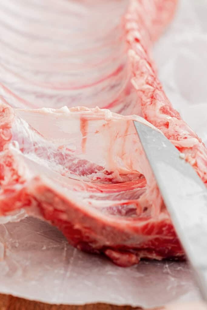 A close-up of a knife removing the membrane from a raw rack of pork ribs on parchment paper for slow cooker garlic honey ribs.