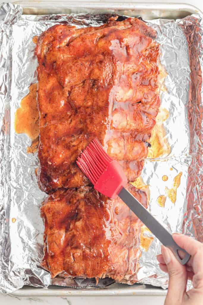 A hand uses a red silicone brush to add sauce to cooked pork ribs on a foil lined pan for slow cooker garlic honey ribs.