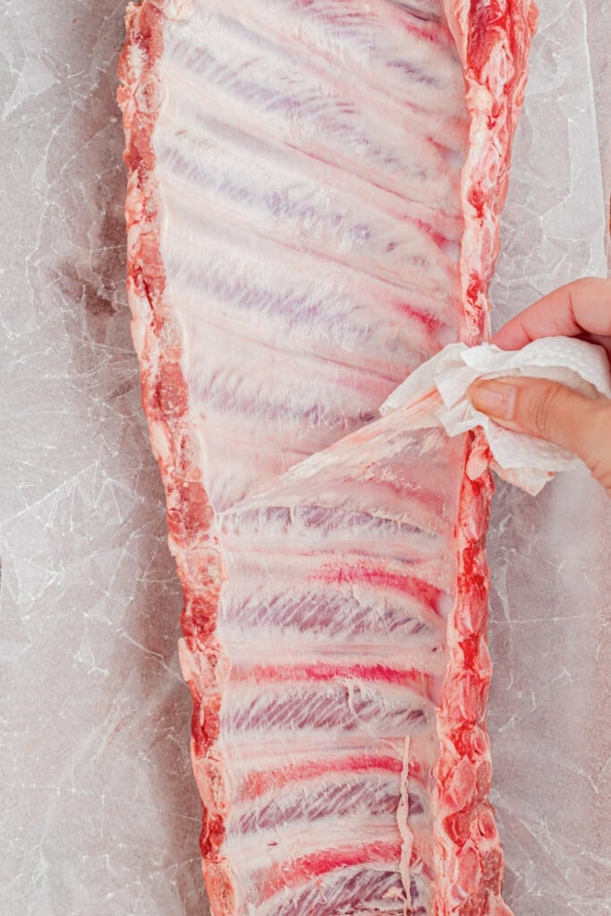 A hand uses a paper towel to peel the membrane off the back of a raw rack of ribs on a sheet of parchment paper for slow cooker garlic honey ribs.