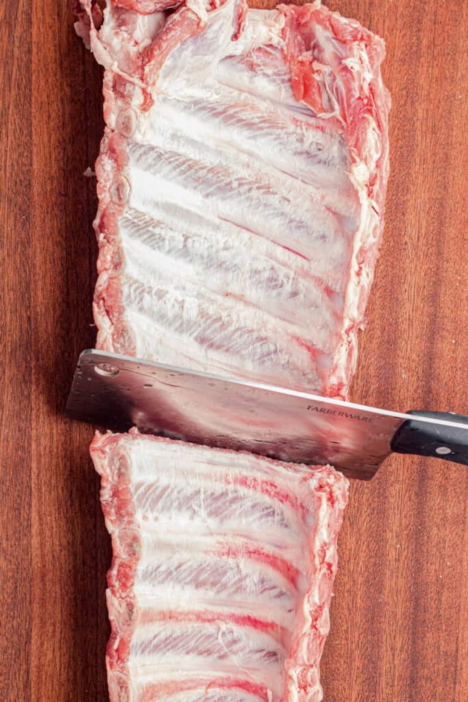 A meat cleaver cutting through a rack of raw pork ribs on a wooden cutting boar for slow cooker honey garlic ribs.