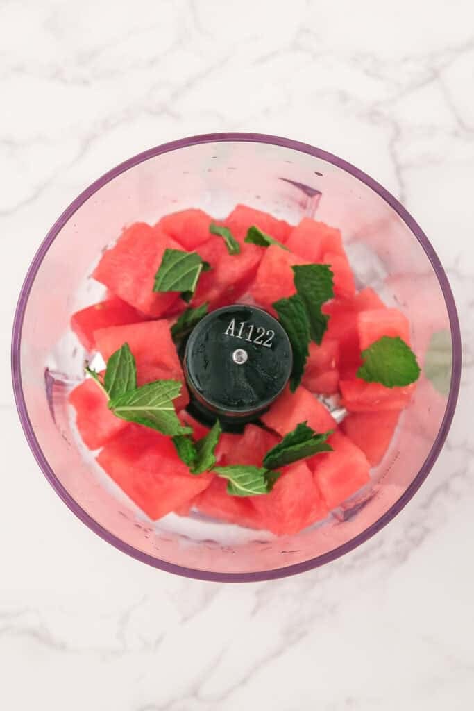 Chopped watermelon and fresh mint leaves in a food processor bowl for watermelon frozen dog treats.