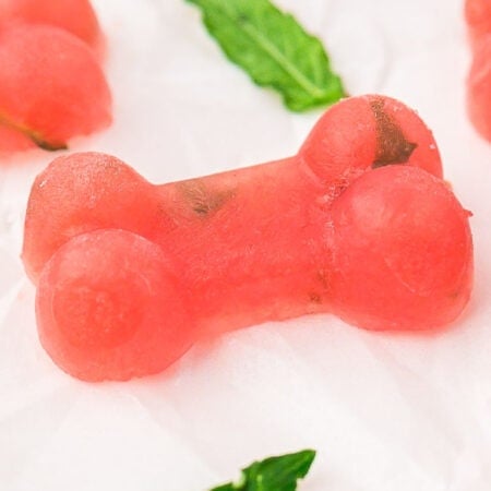 Pink, bone-shaped frozen watermelon dog treats with small green specks on a white surface, garnished with mint leaves.
