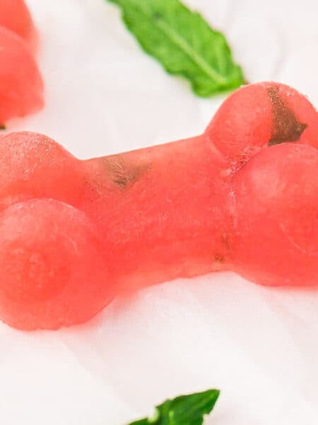 Pink, bone-shaped frozen watermelon dog treats with small green specks on a white surface, garnished with mint leaves.