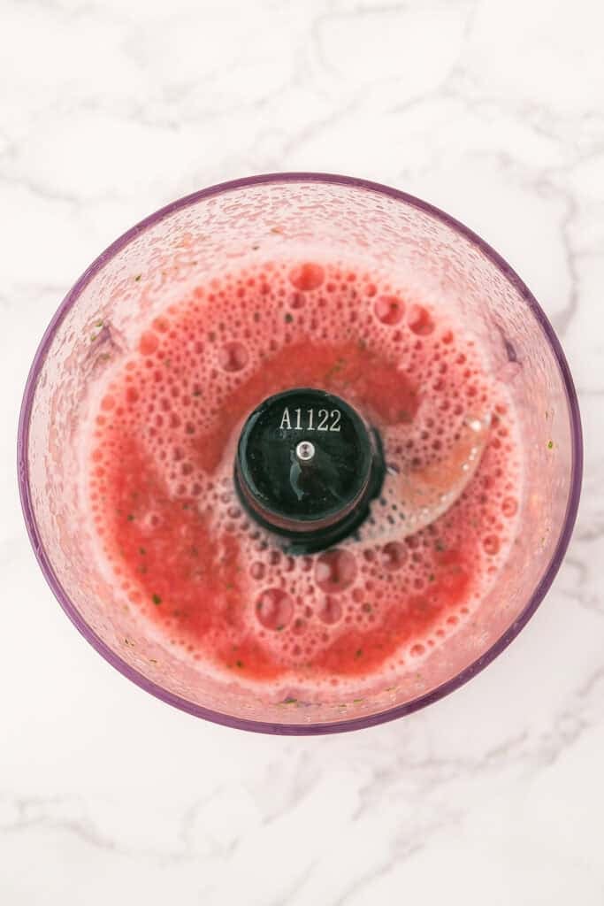 A top-down view of a food processor containing a pink, frothy liquid mixture on a white marble surface.