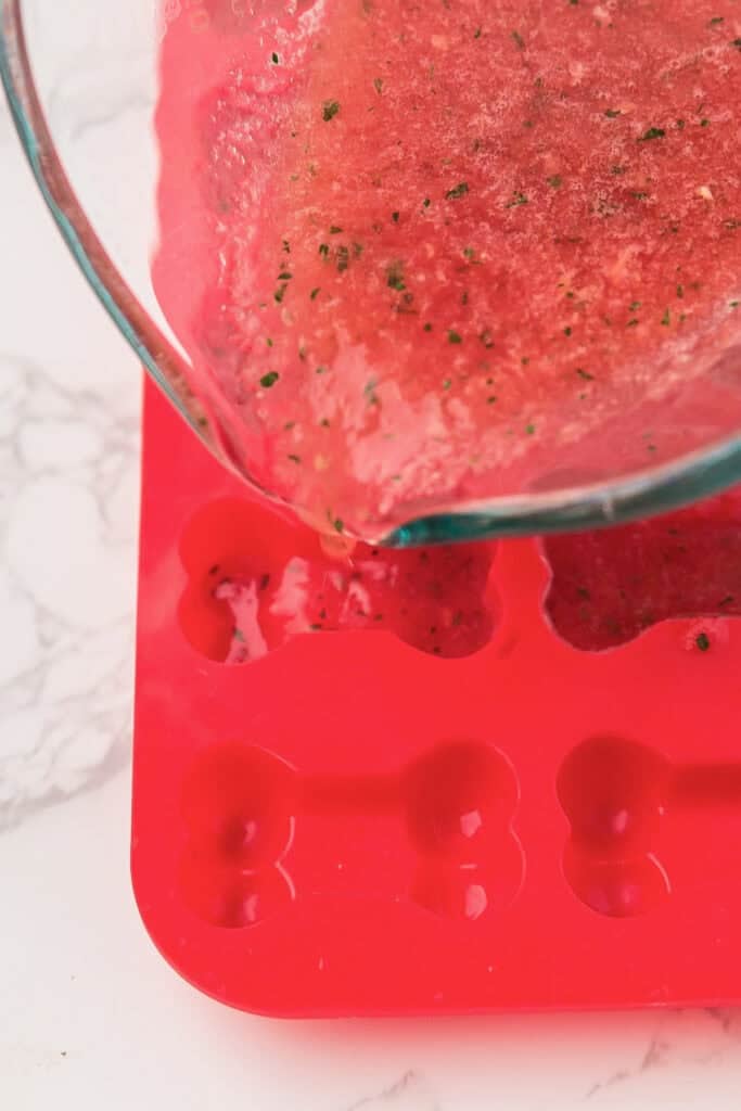 A glass measuring cup pours a watermelon dog treat mixture speckled with mint into a bone mold for watermelon frozen dog treats.