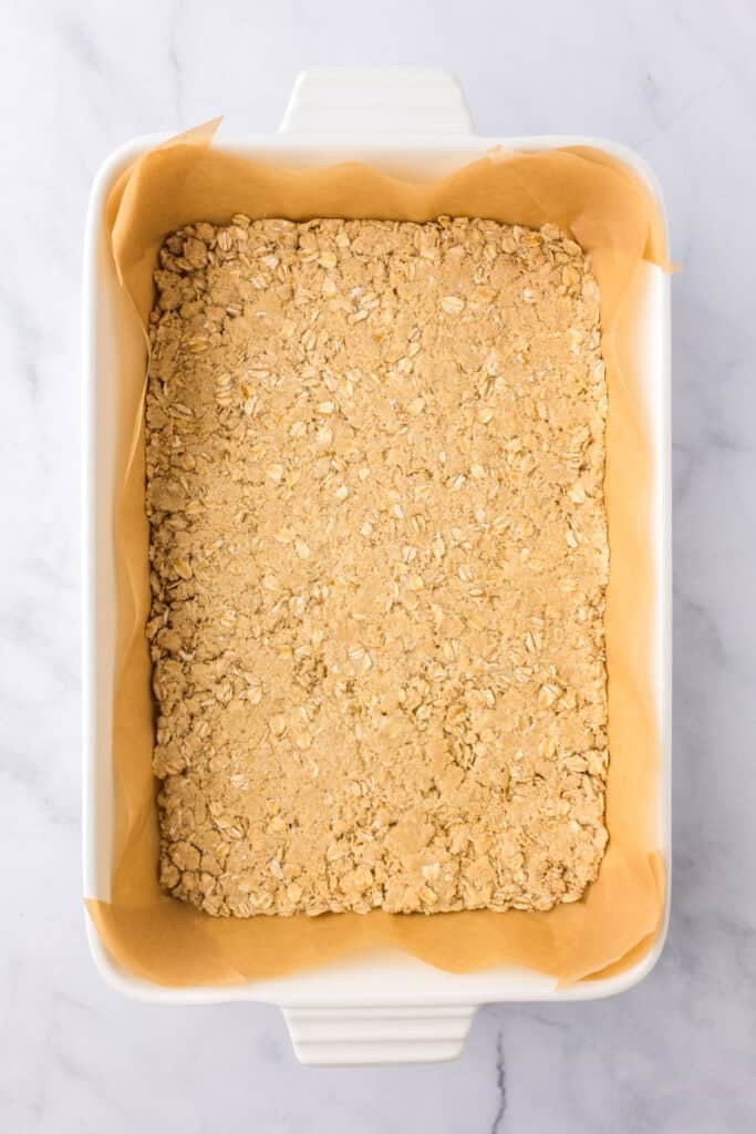 Rectangular white baking dish lined with parchment paper, filled with an even layer of oar crust mixture pressed evenly into the pan for cherry pie bars.