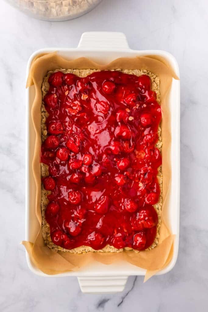 A rectangular baking dish lined with parchment paper with a layer of oat crust and cherry pie filling evenly spread for cherry pie bars.