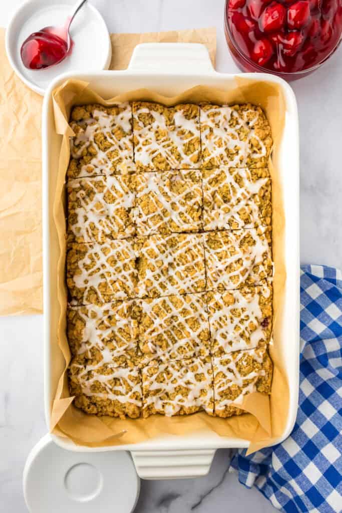A rectangular white baking dish filled with cherry pie bars toped with a crumble topping and a glaze drizzle after slicing.