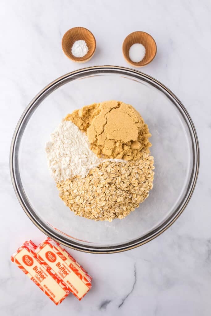 A glass bowl with oats, brown sugar, and flour for the crust for cherry pie bars with butter, salt and baking soda nearby.