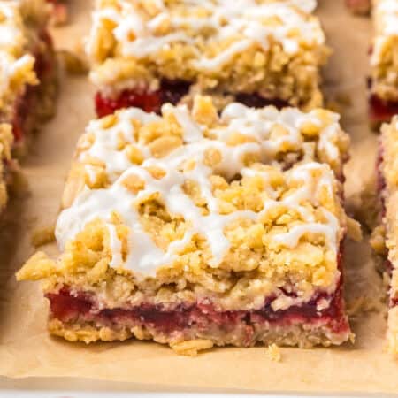 A close-up of a cherry pie bar with a red cherry fruit filling and white icing drizzle, resting on parchment paper.