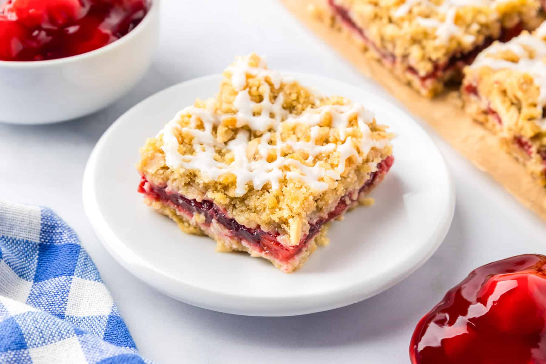 A cherry pie bar bar topped with white icing is served on a white plate, with more bars nearby on the table.