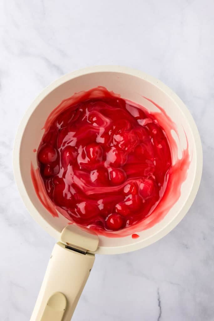 A white saucepan filled with glossy red cherry pie filling sits on a light marble surface.