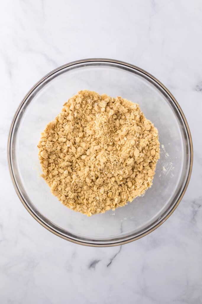 A glass bowl filled with a crumbly mixture of flour, oats and butter for the crust of the cherry pie bars.