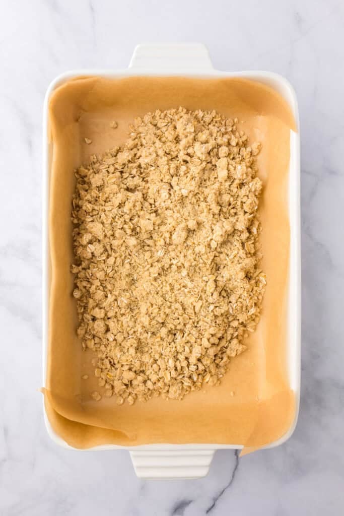 A rectangular baking dish lined with parchment paper is filled with an even layer of crumbly oat crust for cherry pie bars.