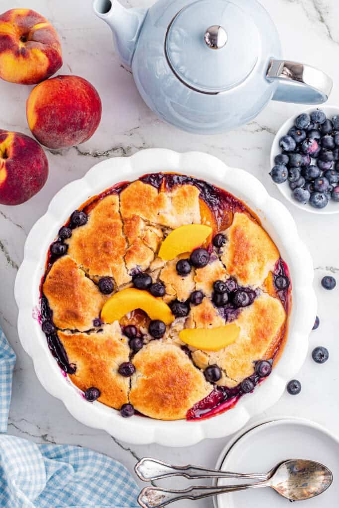 A baked peach and blueberry cobbler in a white dish ready to serve on a table from overhead.
