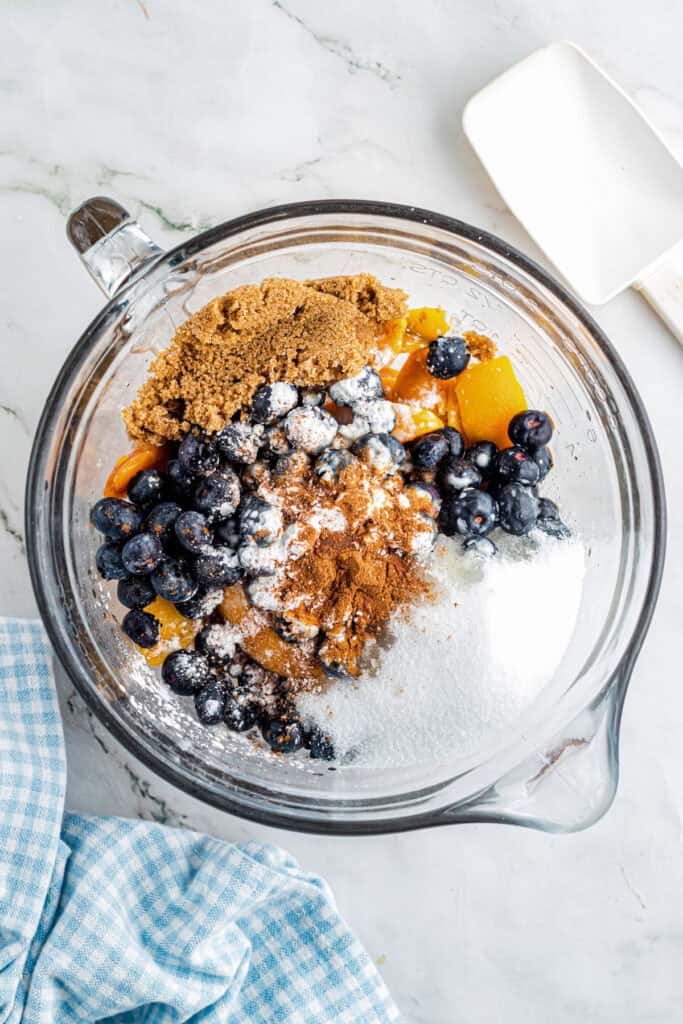 A glass bowl containing brown sugar, white sugar, blueberries, peaches, cinnamon, and flour bring mixed got peach blueberry cobbler.