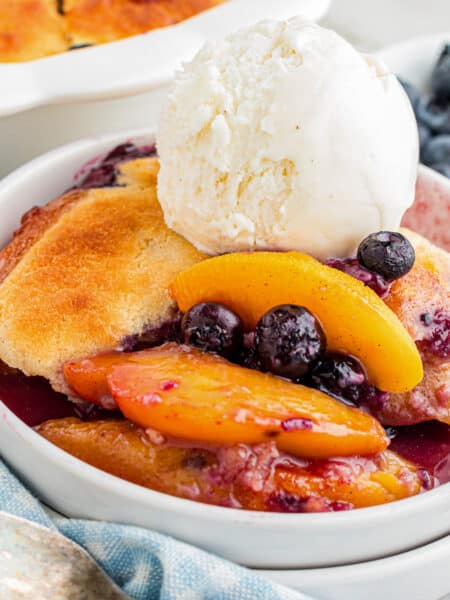 A zoomed in look of a bowl of peach and blueberry cobbler topped with a scoop of vanilla ice cream, with fresh blueberries on the side.
