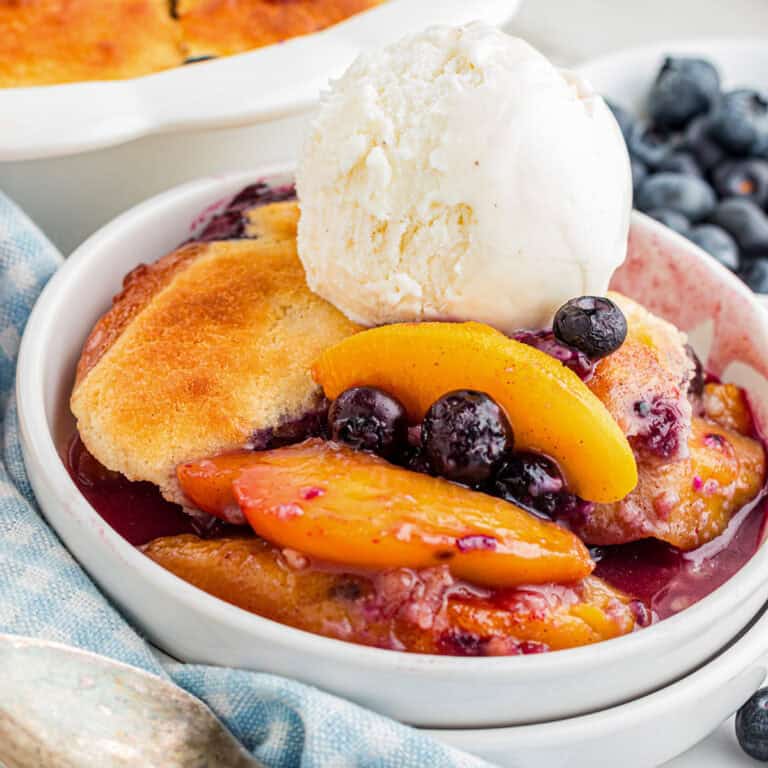 A zoomed in look of a bowl of peach and blueberry cobbler topped with a scoop of vanilla ice cream, with fresh blueberries on the side.