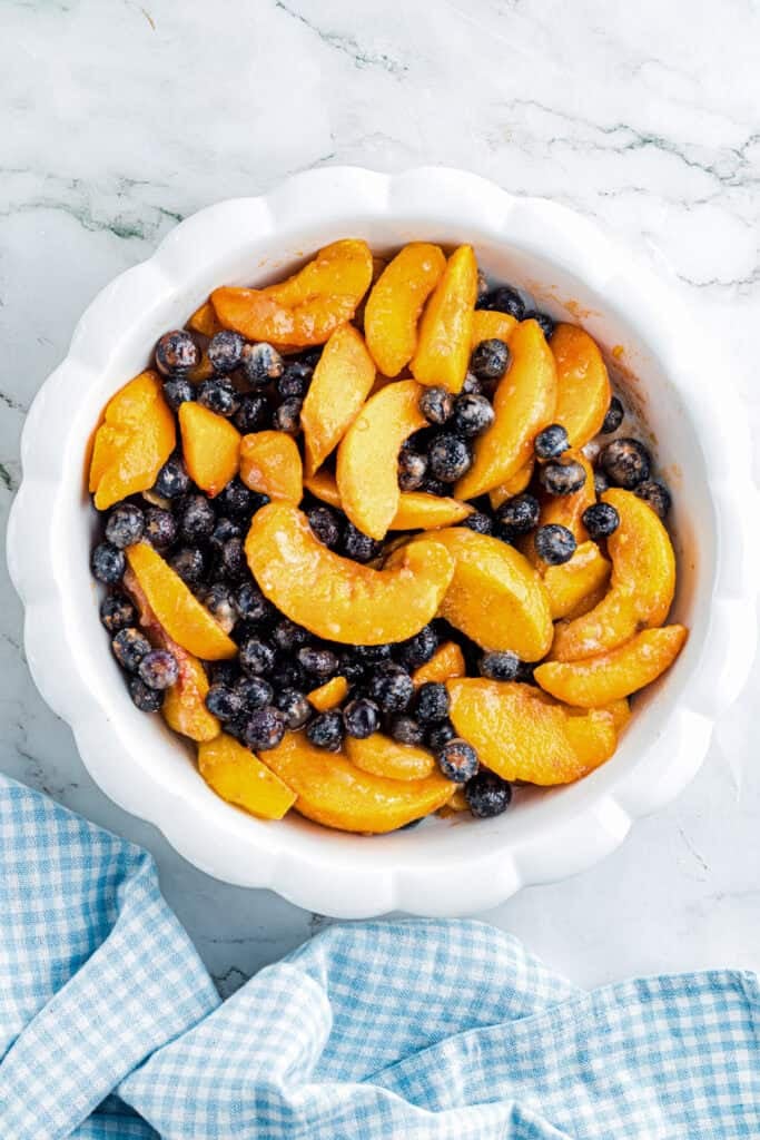 A white pie dish filled with sliced peaches and blueberries and sugar to make peach blueberry cobbler.