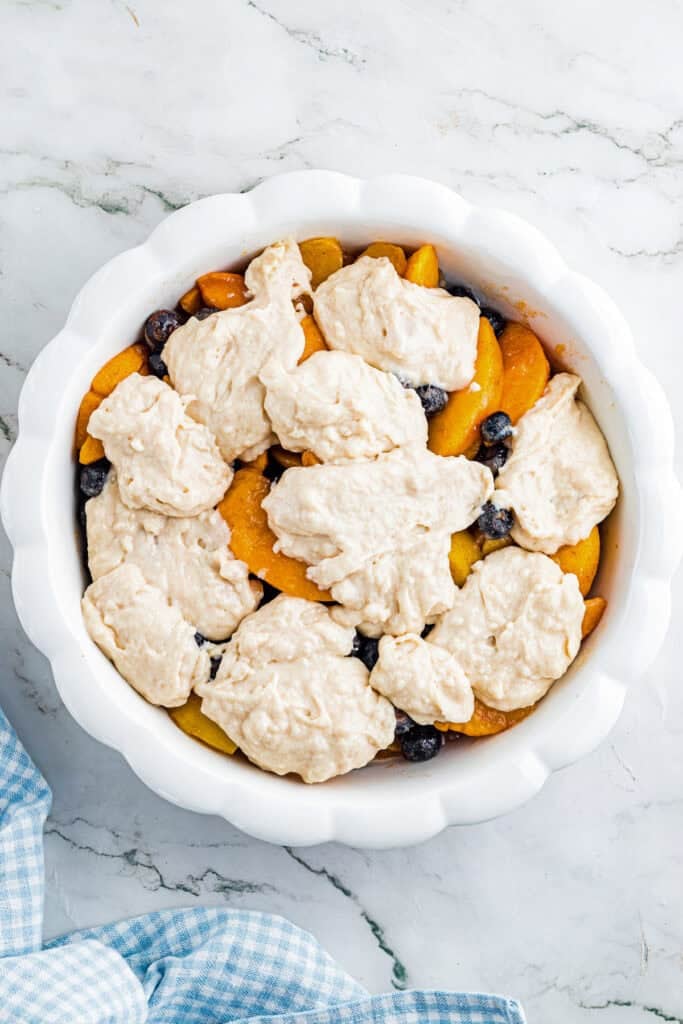 A round baking dish filled with sliced peaches, blueberries, and dollops of unbaked biscuit dough for peach blueberry cobller.