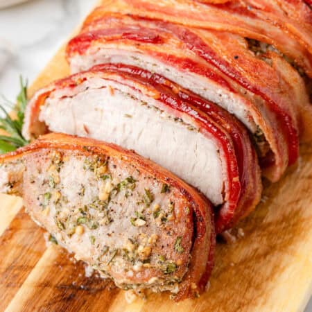 A pork loin wrapped with bacon, sliced on a wooden cutting board, reveals a perfectly seasoned interior and crispy bacon on the outside.