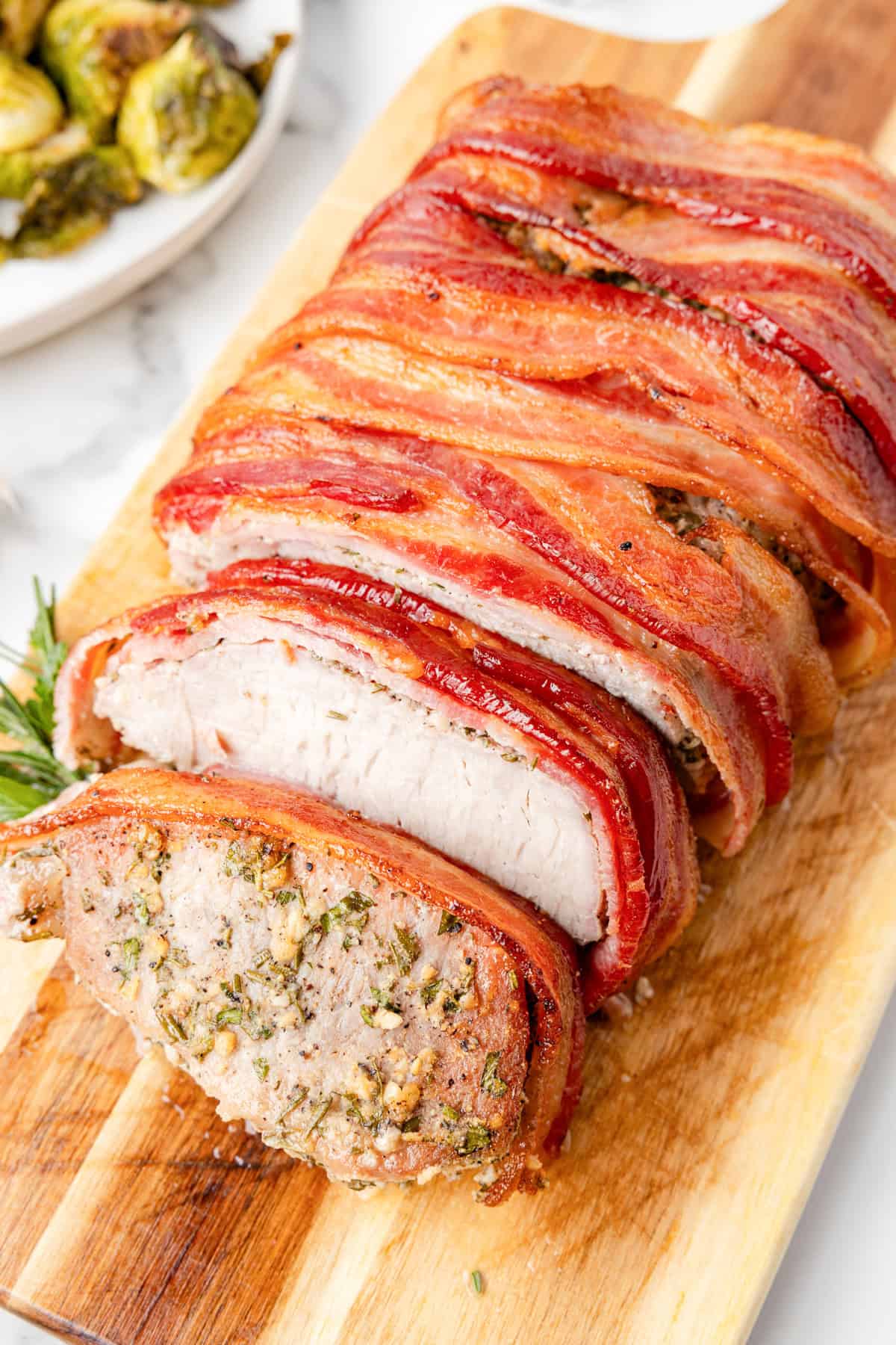 Sliced Pork Loin Wrapped In Bacon on a wooden cutting board, showcasing fragrant herbs tucked inside the tender meat. Roasted Brussels sprouts rest on a plate in the background.