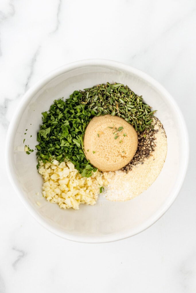 A white bowl containing parsley, rosemary, garlic, brown sugar, black pepper, salt, and onion powder for pork loin wrapped in bacon.