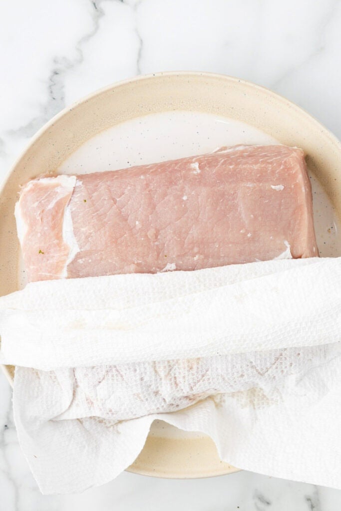 Raw pork loin on a plate with a paper towel patting the meat dry for pork loin wrapped in bacon.