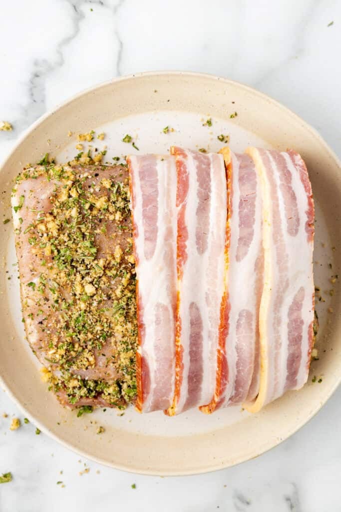 Raw pork loin partially covered with a herb and breadcrumb mixture, and partially wrapped in uncooked bacon for pork loin wrapped in bacon.