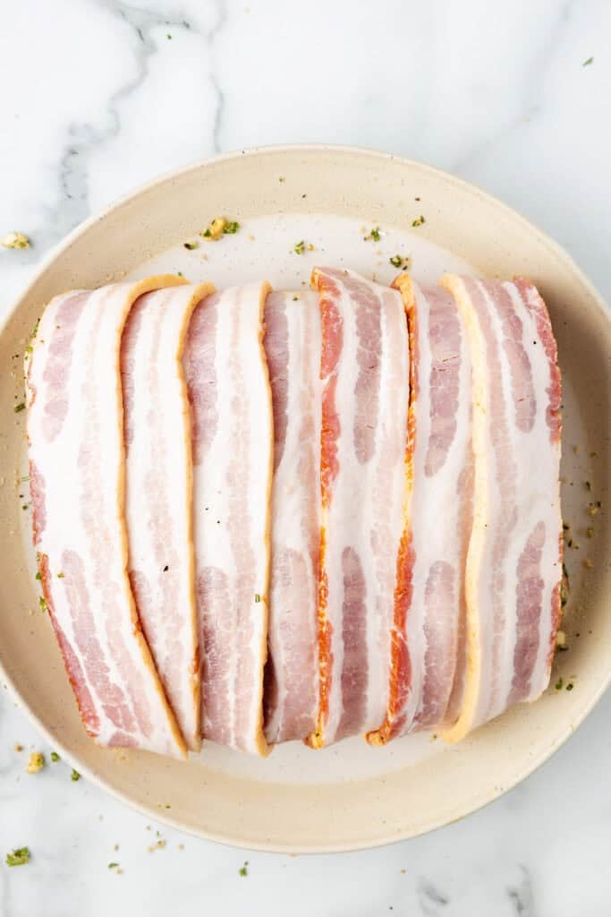 A plate with raw pork loin wrapped in bacon completly with stray herbs and rub on the plate.