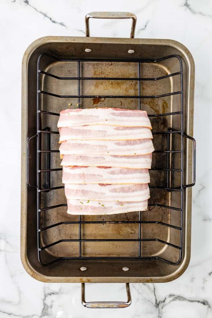 A metal roasting pan with a rack holds a rectangular piece of pork loin covered with evenly spaced strips of raw bacon for pork loin wrapped in bacon.