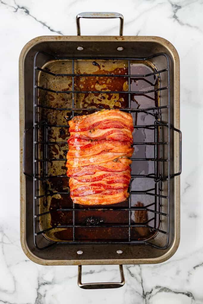 Rectangular pork loin wrapped in bacon strips sits on a rack in a metal roasting pan, with meat juices pooled at the bottom.