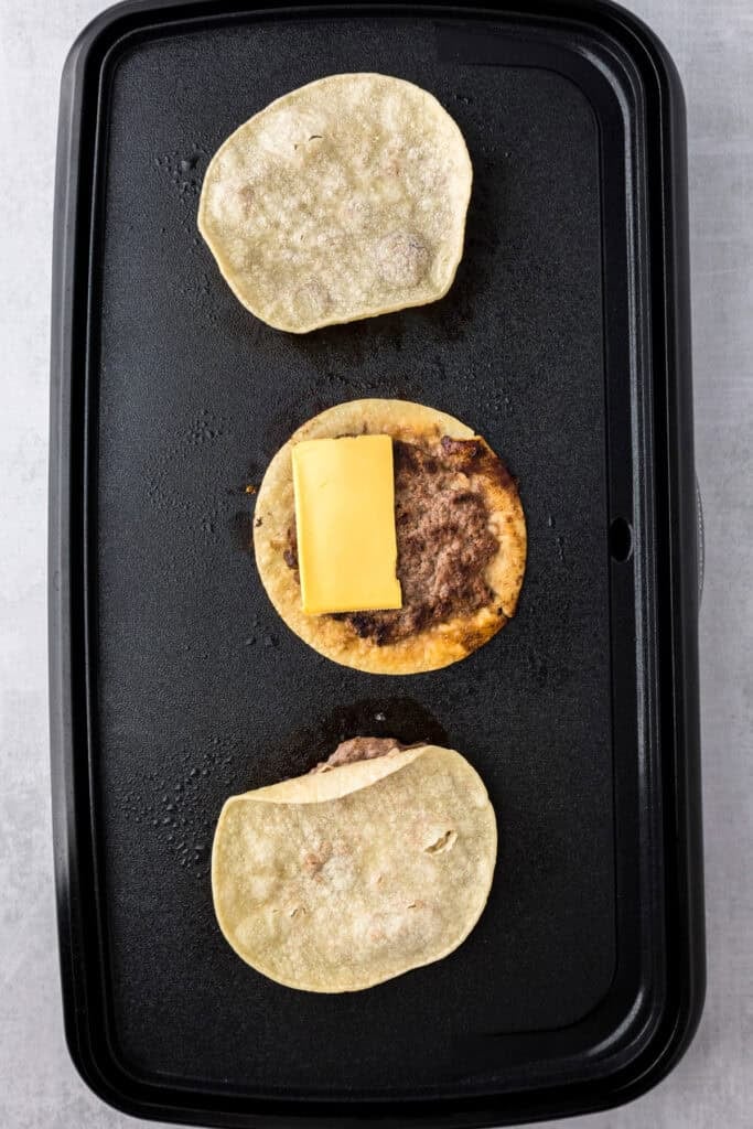 Three corn tortillas on a griddle, the middle one with ground beef and a slice of cheese, while the top and bottom tortillas meat side down for burger smash tacos.