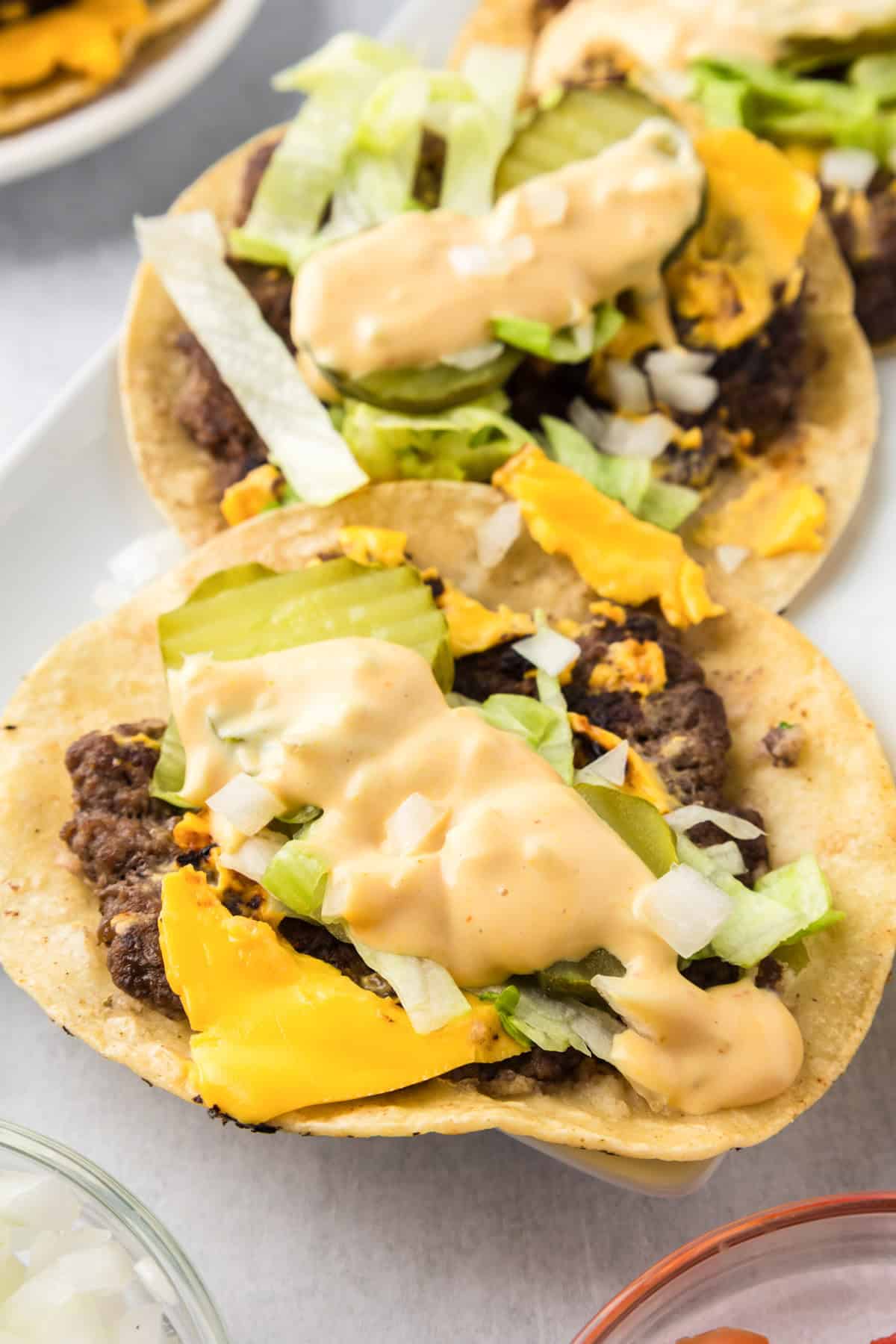 Two open-faced smash burger tacos with ground beef, melted cheese, chopped onions, lettuce, pickles, and creamy sauce on a tortilla.