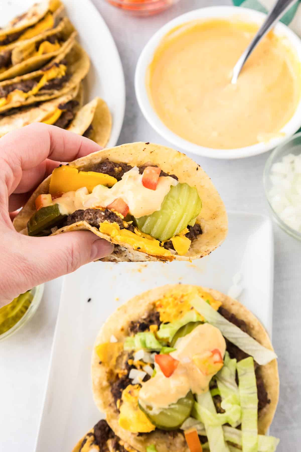A hand holds a smash burger taco filled with ground beef, cheese, pickles, and sauce. Other assembled tacos and a bowl of sauce are on the table.