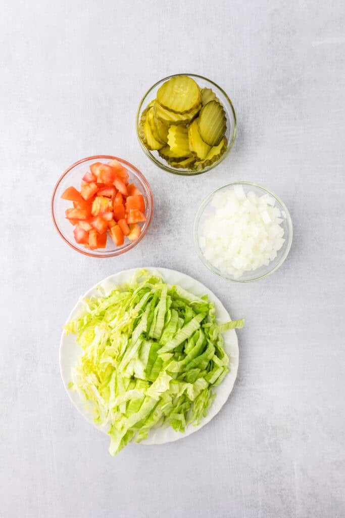 Four bowls containing sliced pickles, diced tomatoes, diced onions, and shredded lettuce as toppings for smash burger tacos.