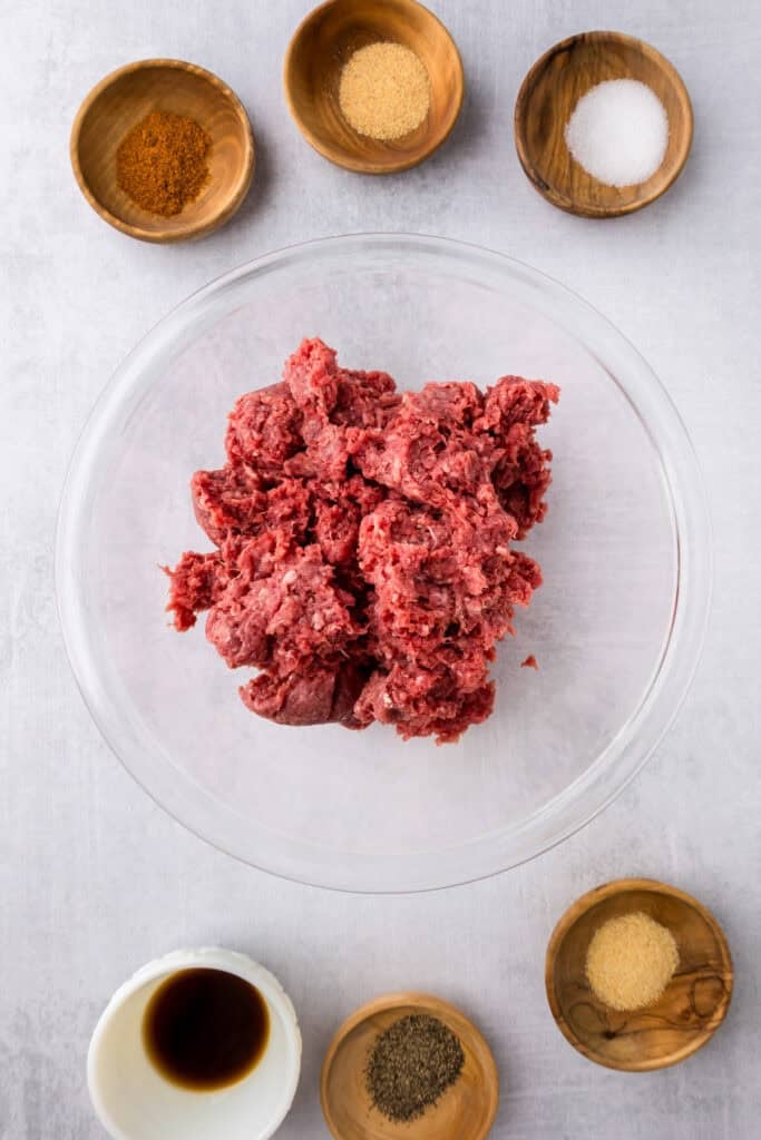 A glass bowl of raw ground beef is surrounded by small bowls containing various spices, salt, pepper, and a small bowl of Worcestershire sauce for smash burger tacos.