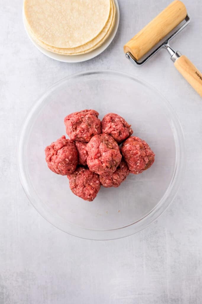 A clear bowl with raw ground beef for burger smash tacos formed into balls, next to a plate of tortillas and a small rolling pin.
