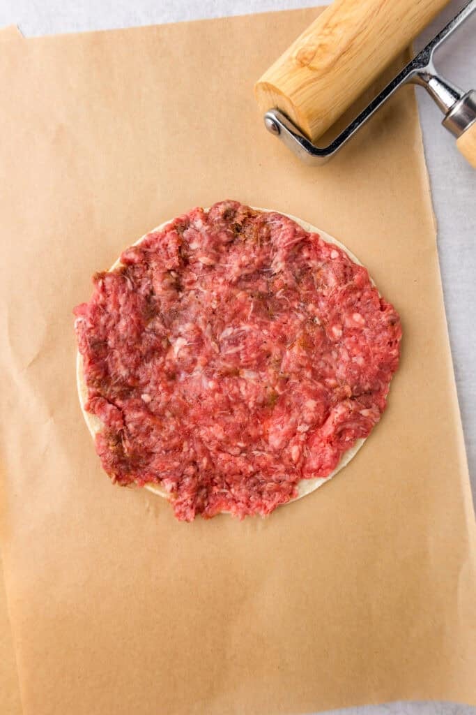 Raw ground meat spread in a thin layer on a corn tortilla for smash burger tacos placed on parchment paper next to a wooden rolling pin.